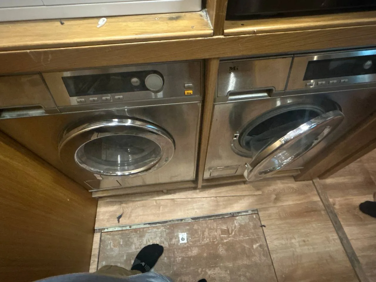 A pair of stainless steel washing machines, one with the door open, with a technician's feet visible at the bottom of the
