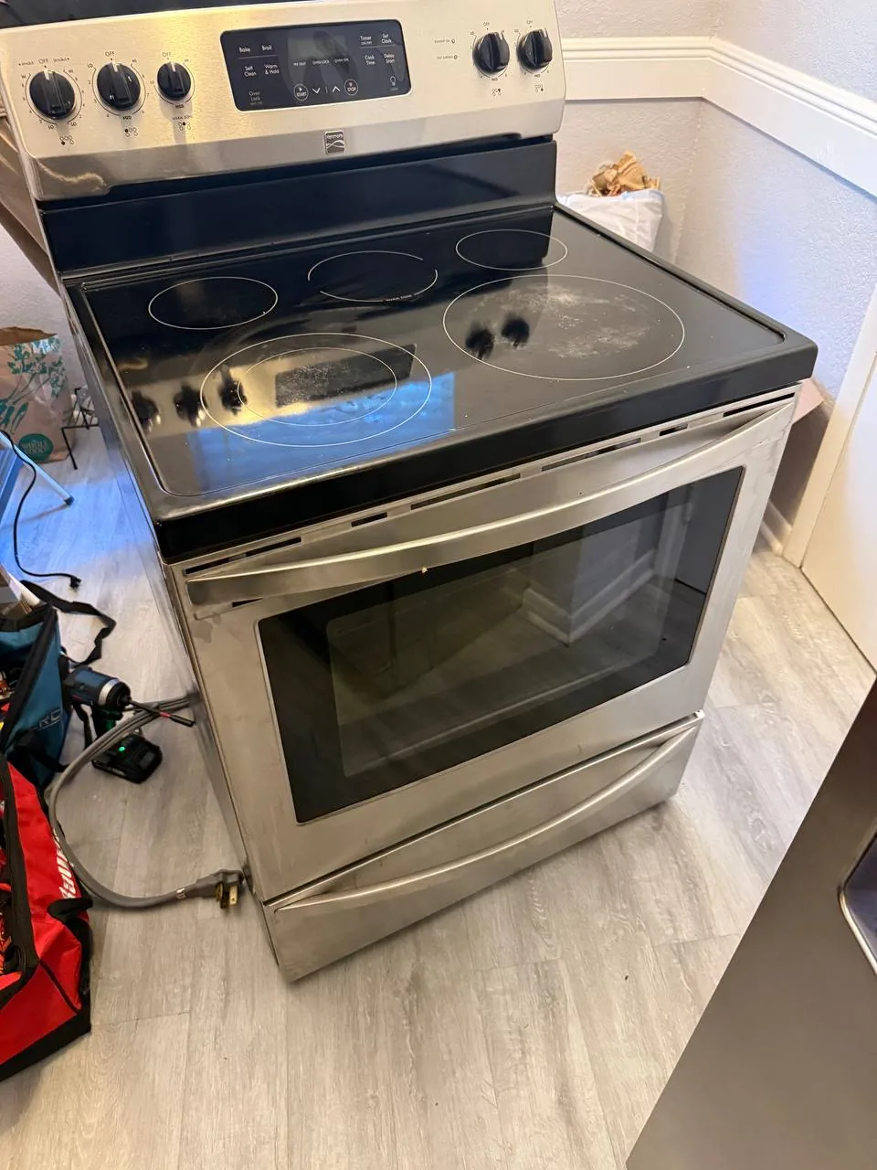 A stainless steel oven with a black electric cooktop, pulled out from its spot with the power cord visible on the floor