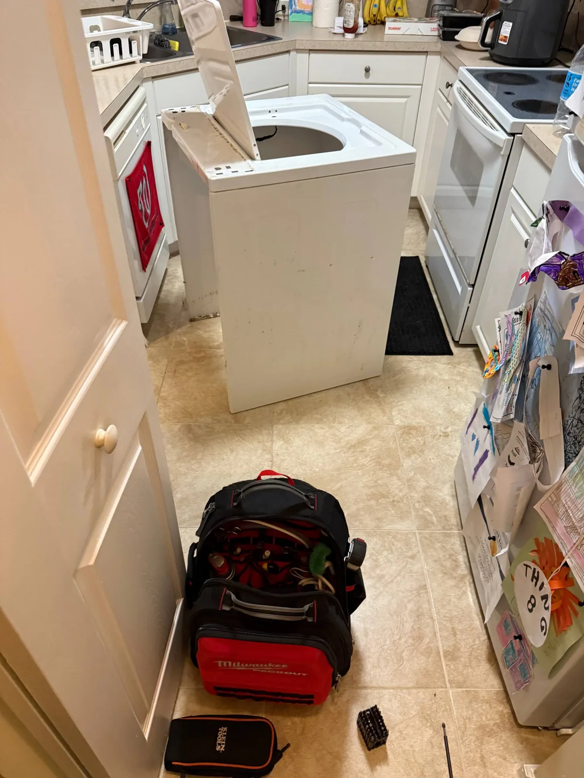 A washer with its top removed sits in a kitchen with a toolbox and various tools on the floor nearby