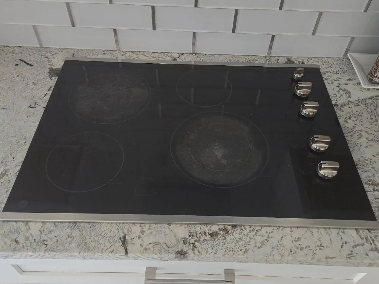 A clean black electric cooktop with four burners and five silver control knobs installed in a speckled granite countertop