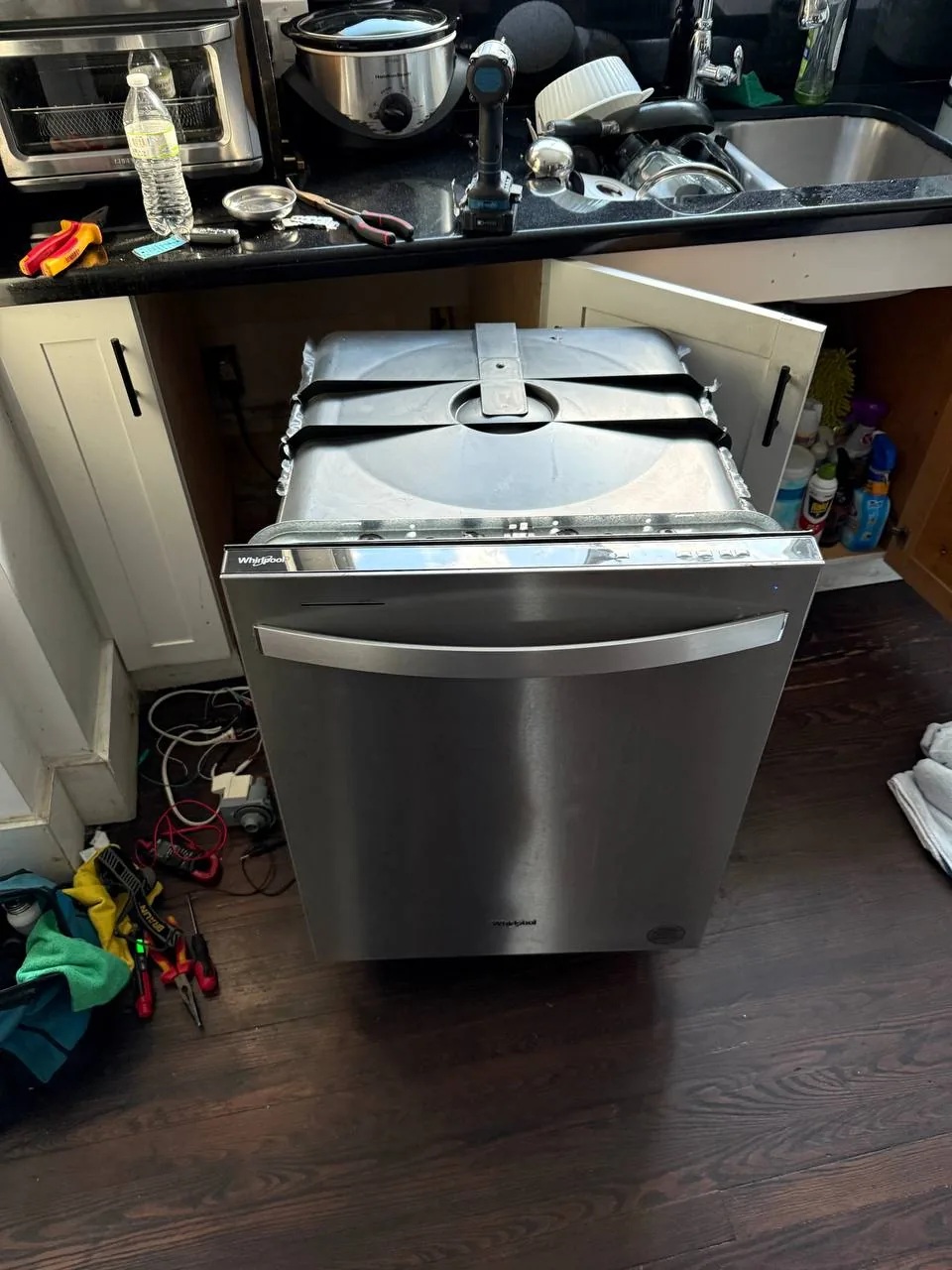 A stainless steel dishwasher is pulled out from under a counter with tools nearby, indicating a repair is underway