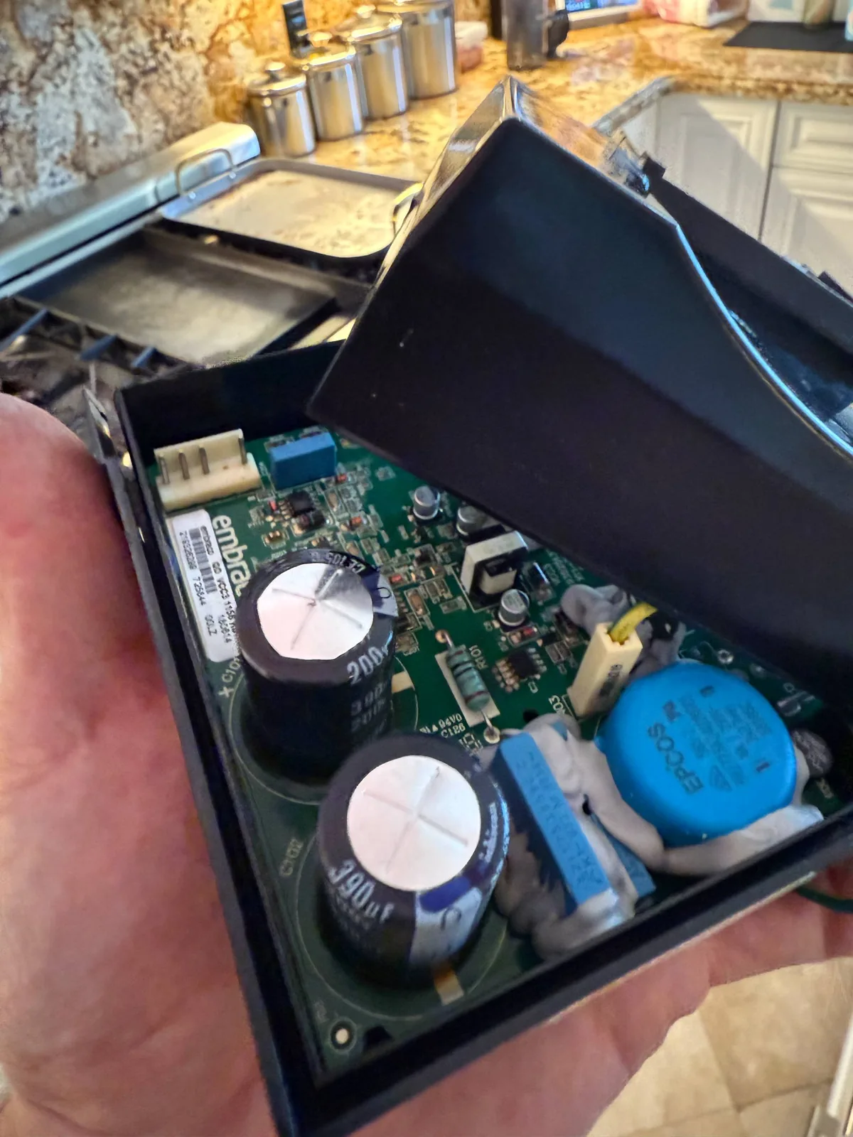 A technician holds an electronic board with capacitors, possibly from a cooktop, with a kitchen countertop in the background