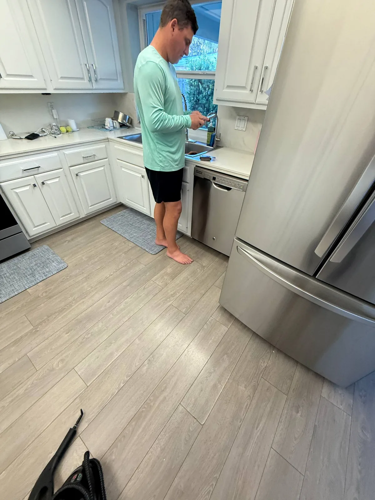 A technician is standing in a kitchen with a stainless steel refrigerator and dishwasher visible, looking at his phone