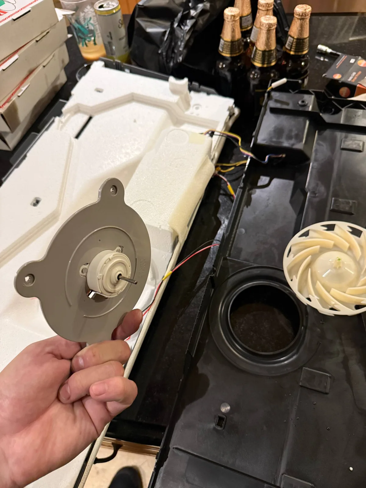 A hand holds a refrigerator fan motor and parts are laid out on a counter during a repair