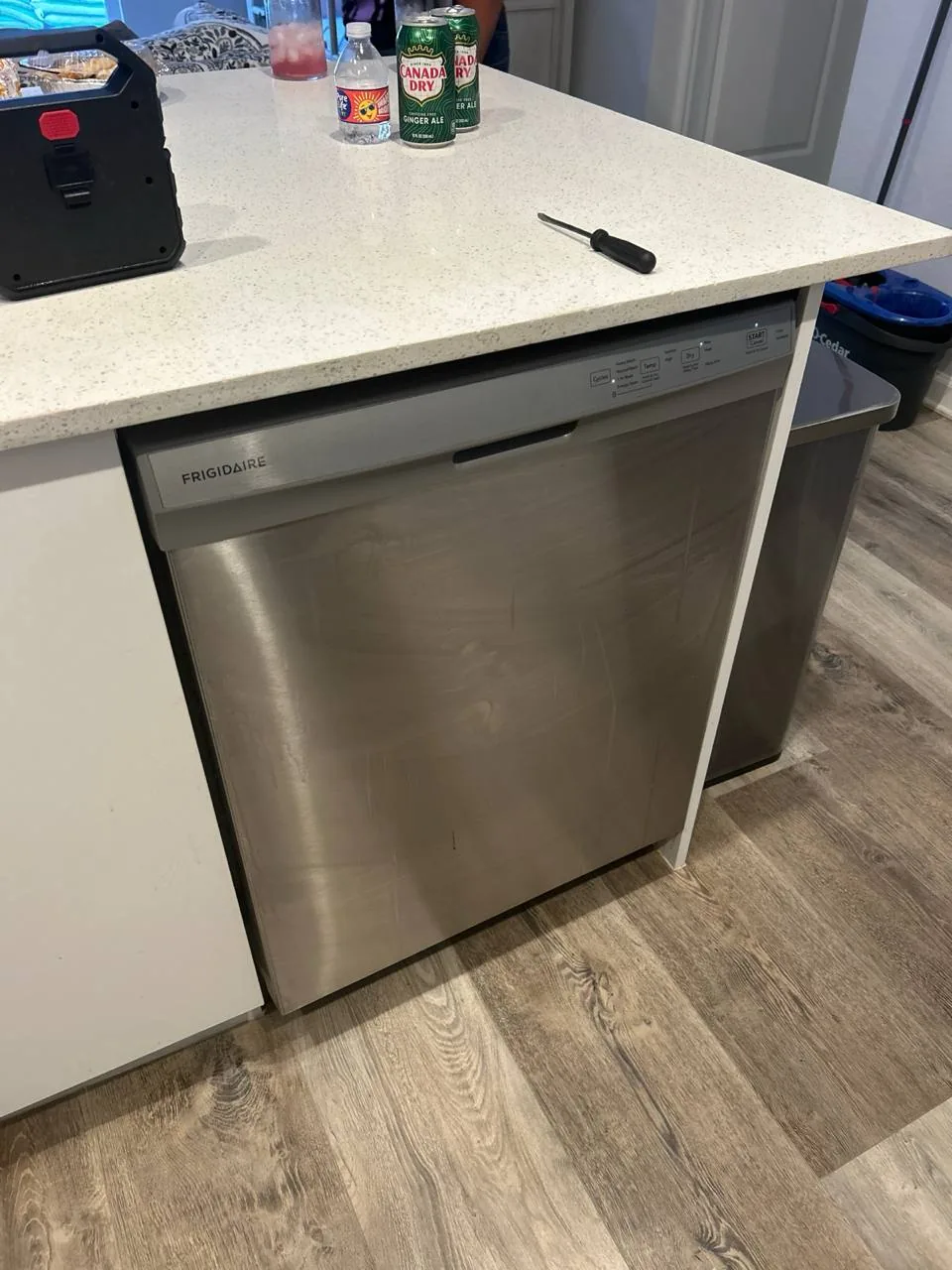 A stainless steel Frigidaire dishwasher is installed under a white countertop, with a screwdriver and drinks on the counter