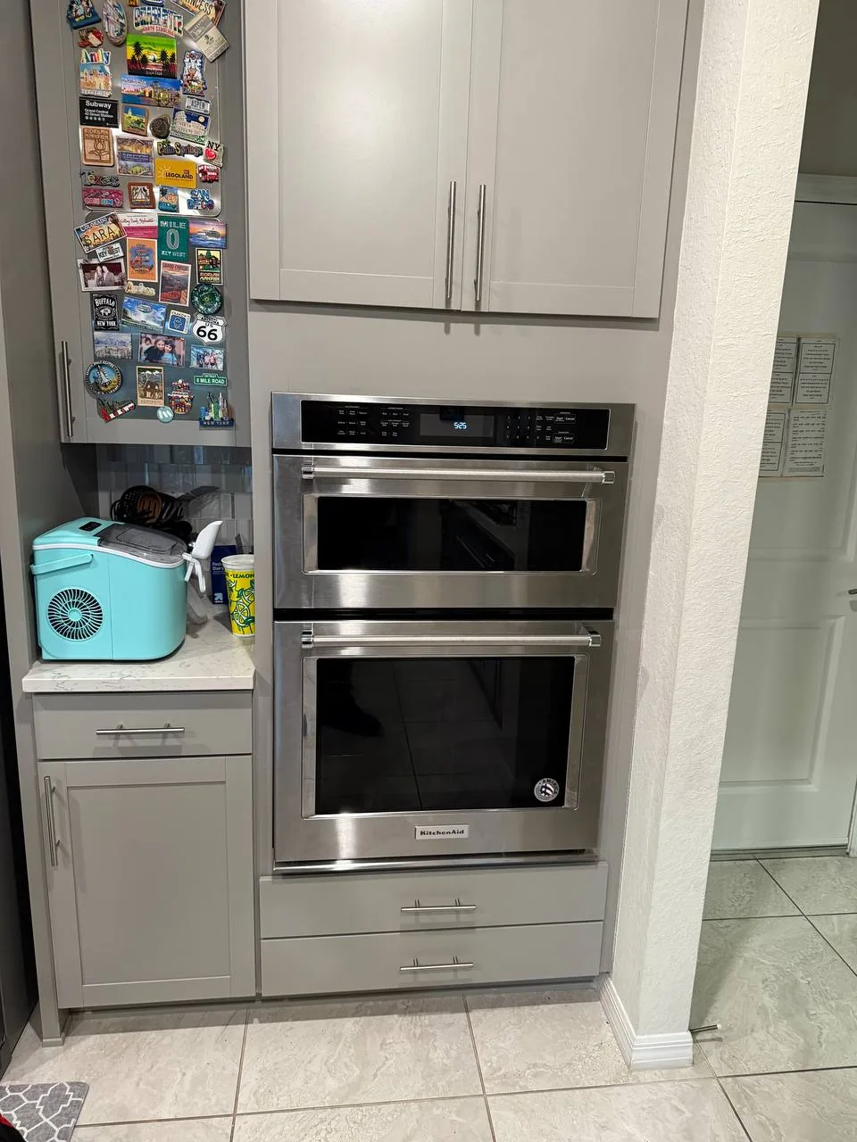 A stainless steel double oven installed in kitchen cabinetry, with a magnet-covered refrigerator visible to the left
