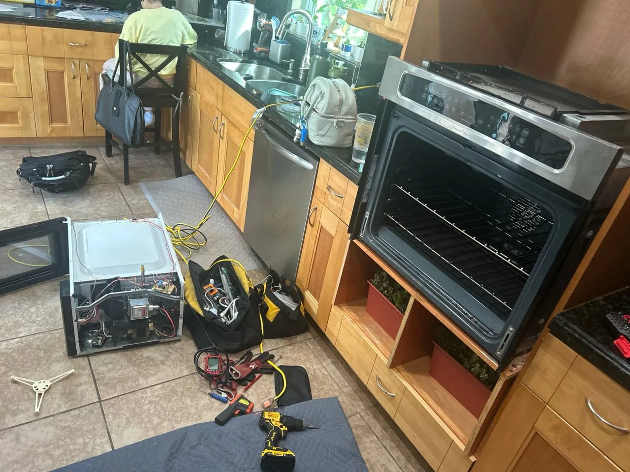 A kitchen scene showing a built-in oven pulled out for repair, with tools and a disassembled microwave on the floor