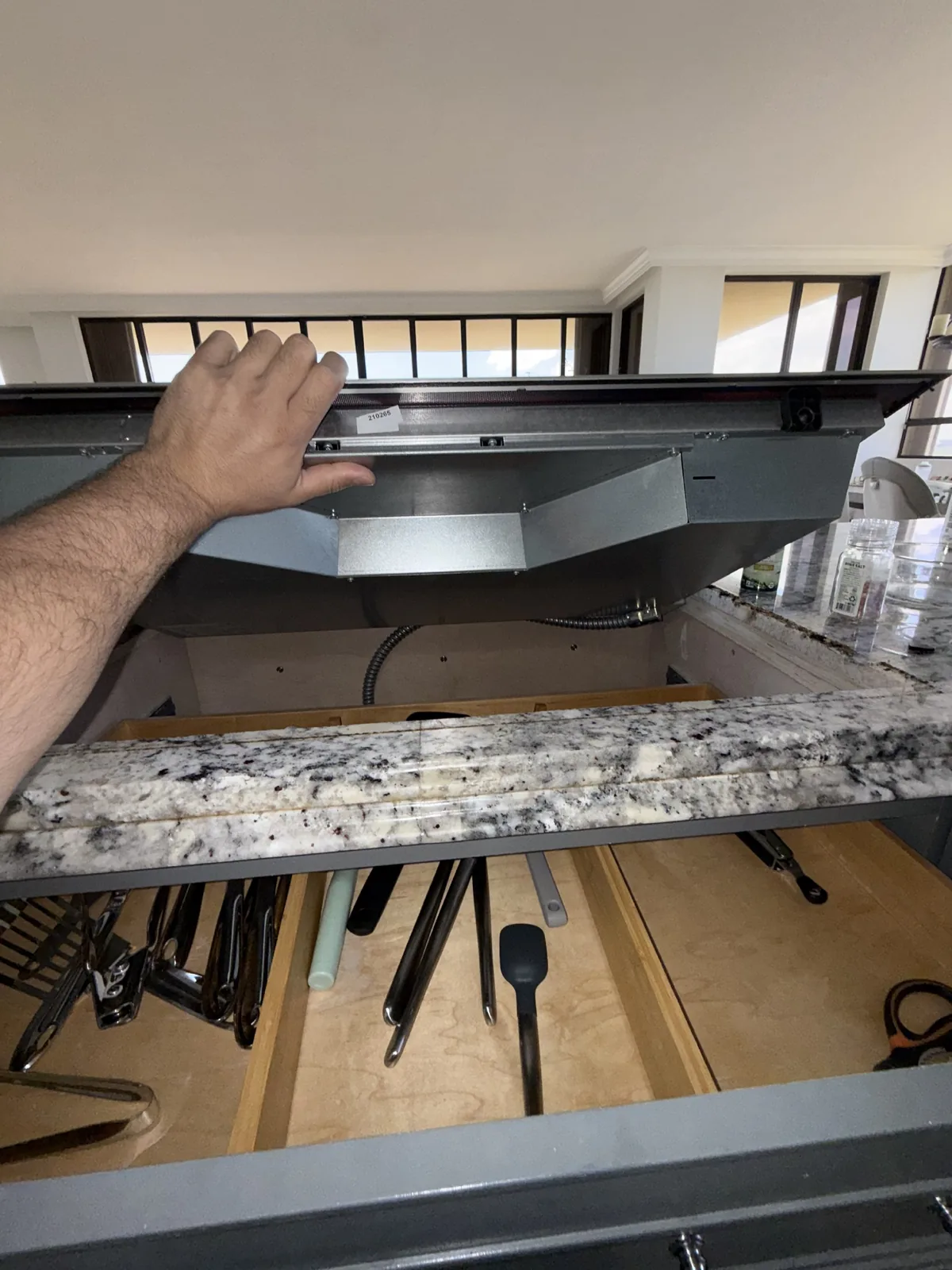 A technician's arm and hand adjusting a cooktop with kitchen utensils in a drawer below