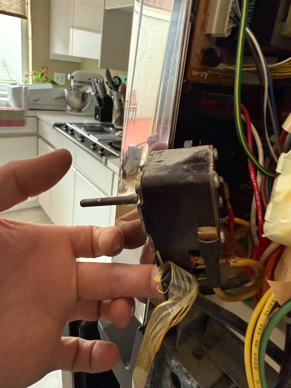 A technician's hand pointing at wiring inside an oven, with a kitchen counter and stovetop in the background