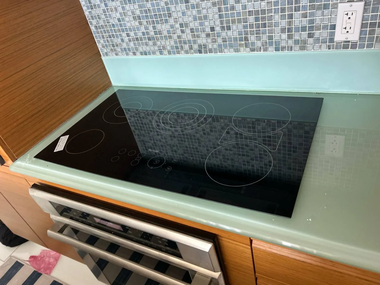 A black glass cooktop is installed in a kitchen counter with light green countertop and blue tiled backsplash
