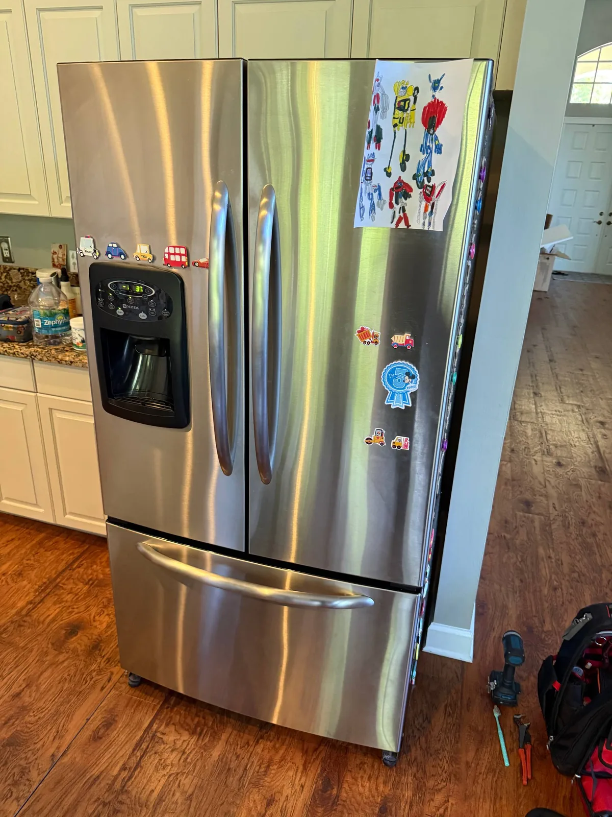 A stainless steel refrigerator with magnets and children's drawings on its doors, with tools on the floor nearby
