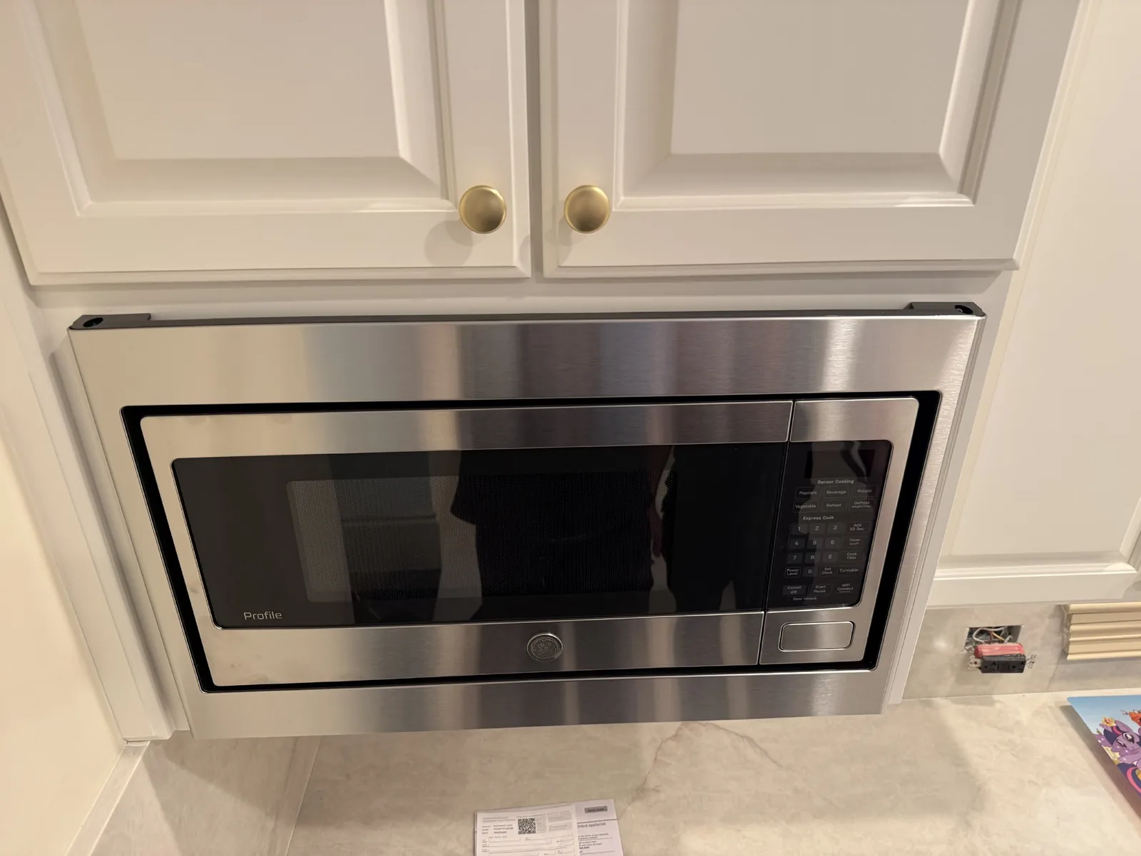 A stainless steel GE Profile microwave installed under white cabinets, with a power outlet visible to the right