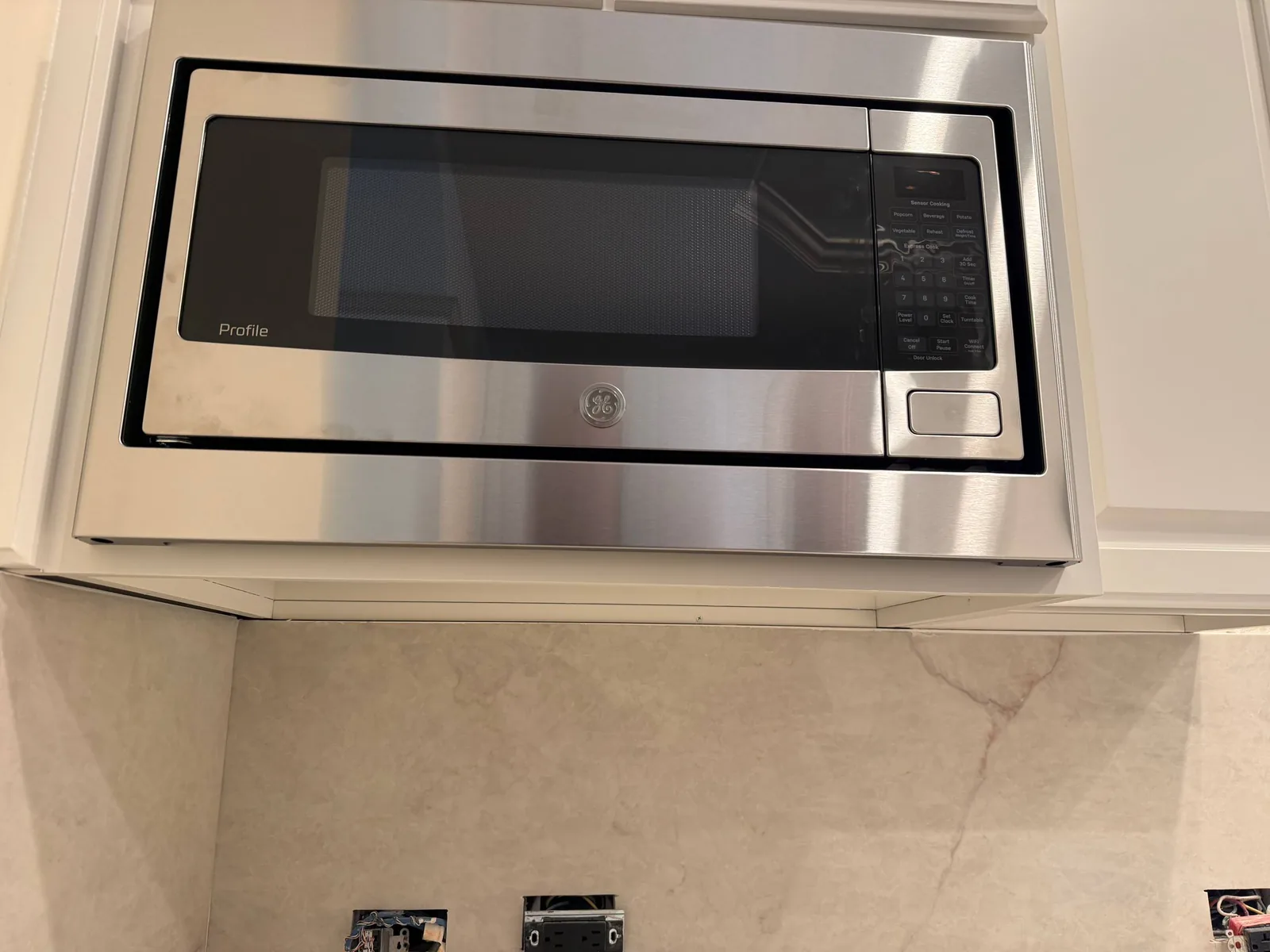 A stainless steel GE Profile microwave installed above a countertop with unfinished electrical outlets below