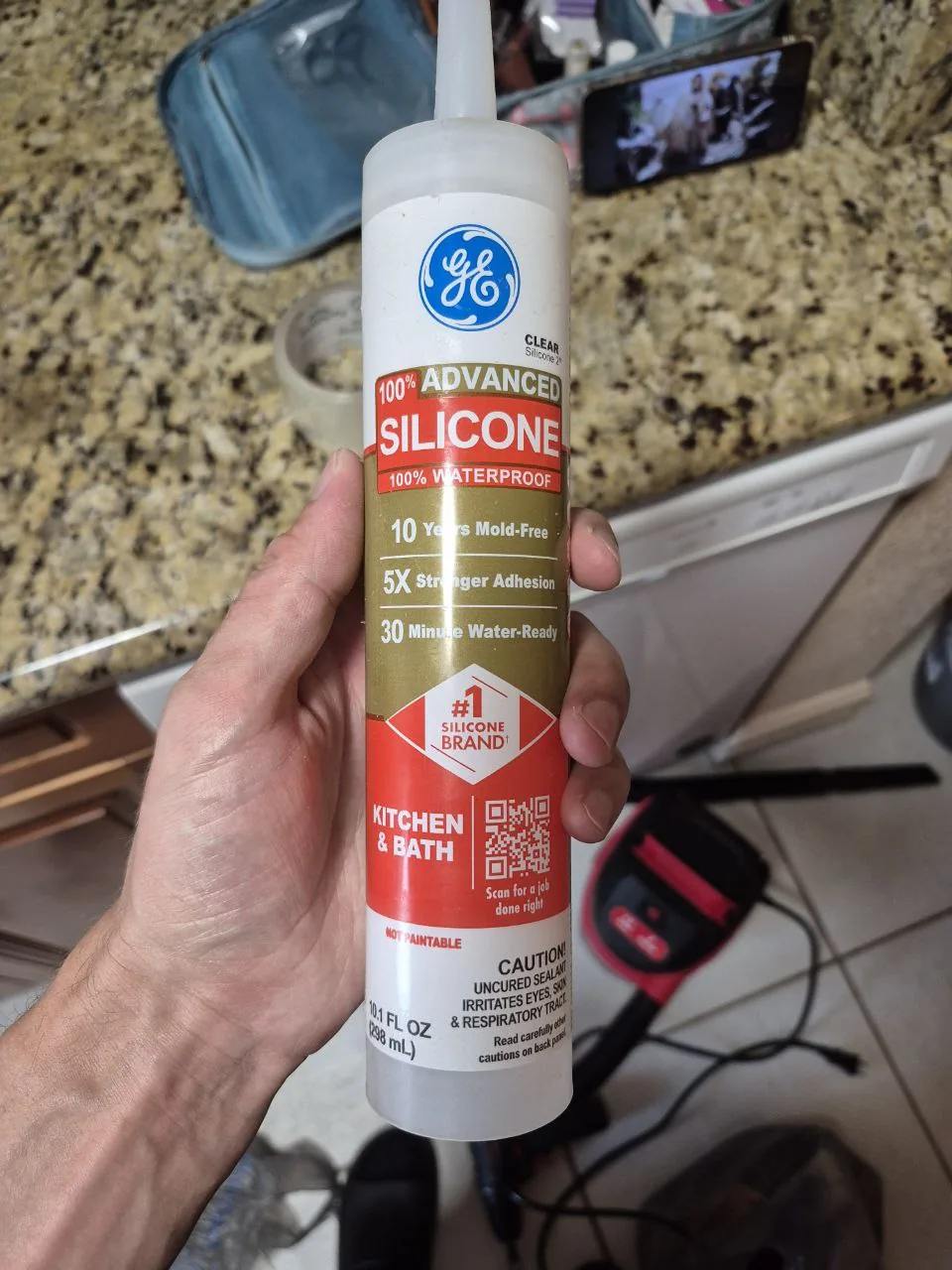 A hand holds a tube of GE Advanced Silicone sealant, with a speckled countertop and tools in the background