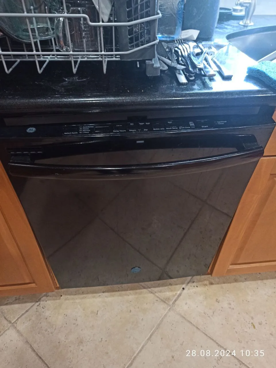 A black GE dishwasher is installed under a black countertop, with some dishes drying in a rack above it