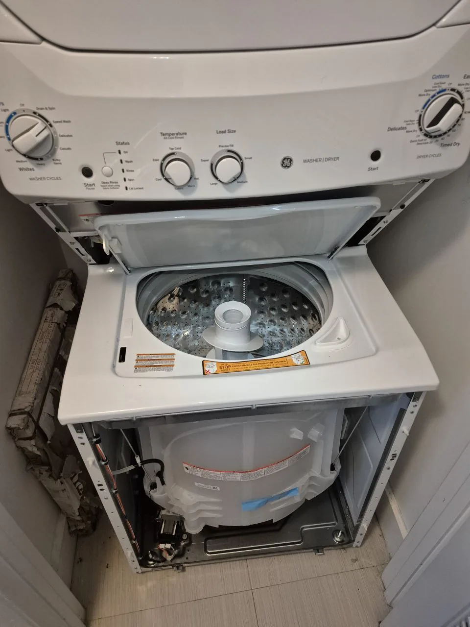 A stacked GE washer and dryer unit with the washer's top lid open and side panel removed, revealing internal components