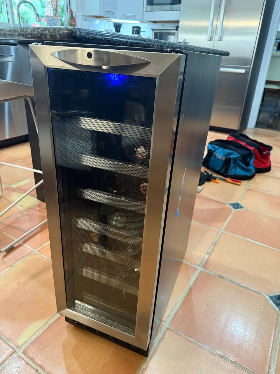 A stainless steel wine cooler with bottles inside in a kitchen setting with tools on the floor