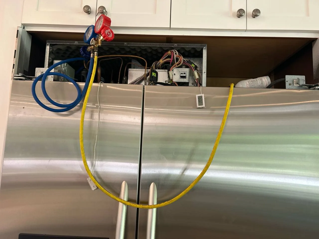 A stainless steel refrigerator with the top panel removed, revealing internal components and repair tools connected