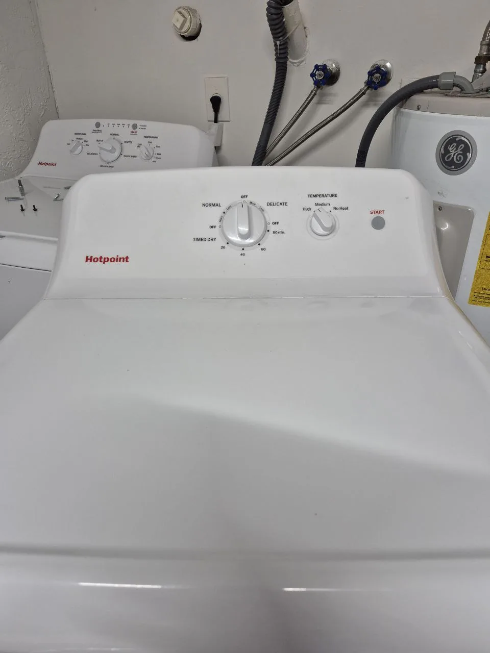 A white Hotpoint dryer with its control panel in focus, next to a matching washer and water heater connections