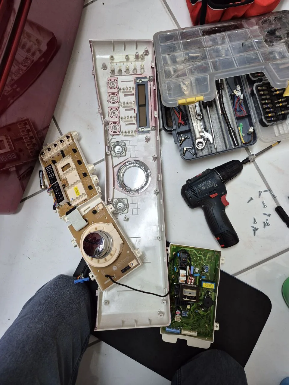 A disassembled washing machine control panel and circuit board are laid on the floor with tools, and a technician's legs