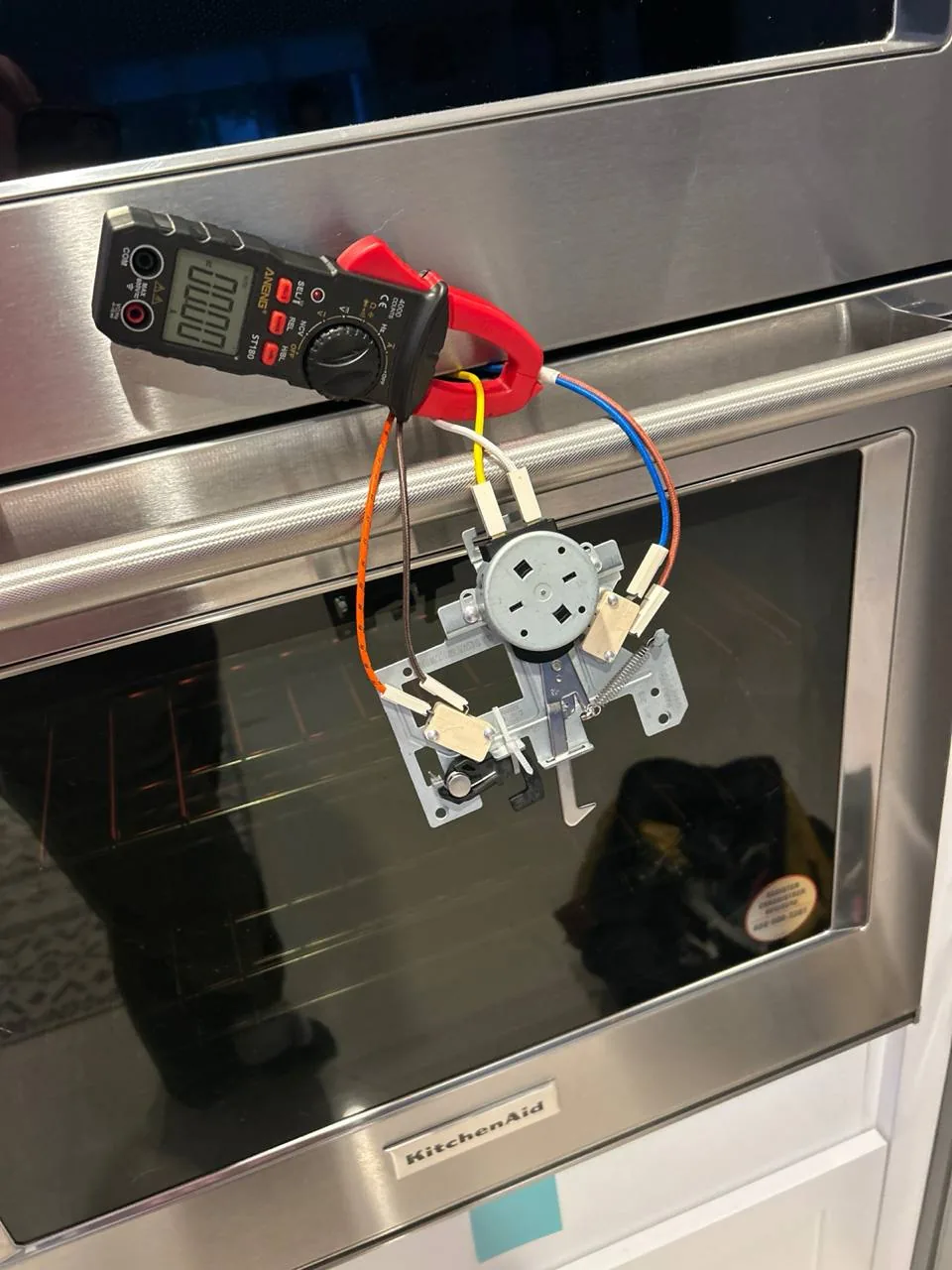 A multimeter is connected to a component in front of a KitchenAid oven