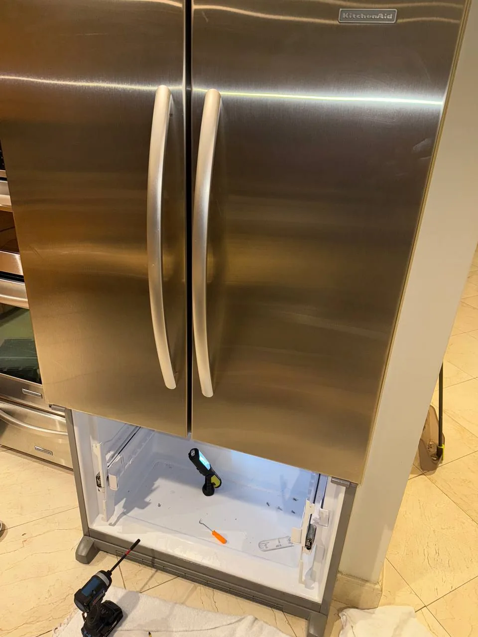A stainless steel KitchenAid refrigerator with its freezer drawer open and repair tools visible