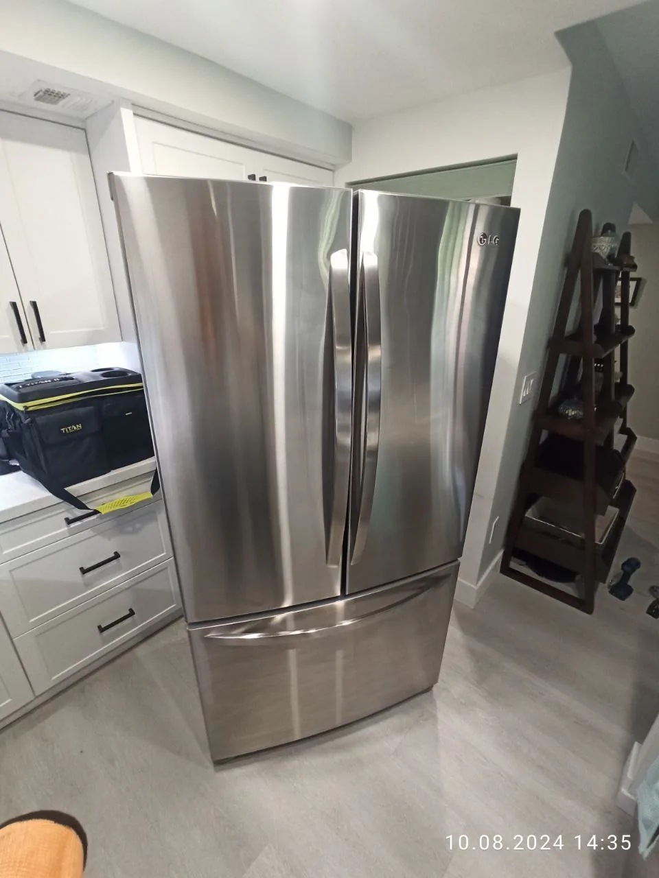 A stainless steel LG French door refrigerator in a kitchen setting with white cabinets and a black tool bag on the counter