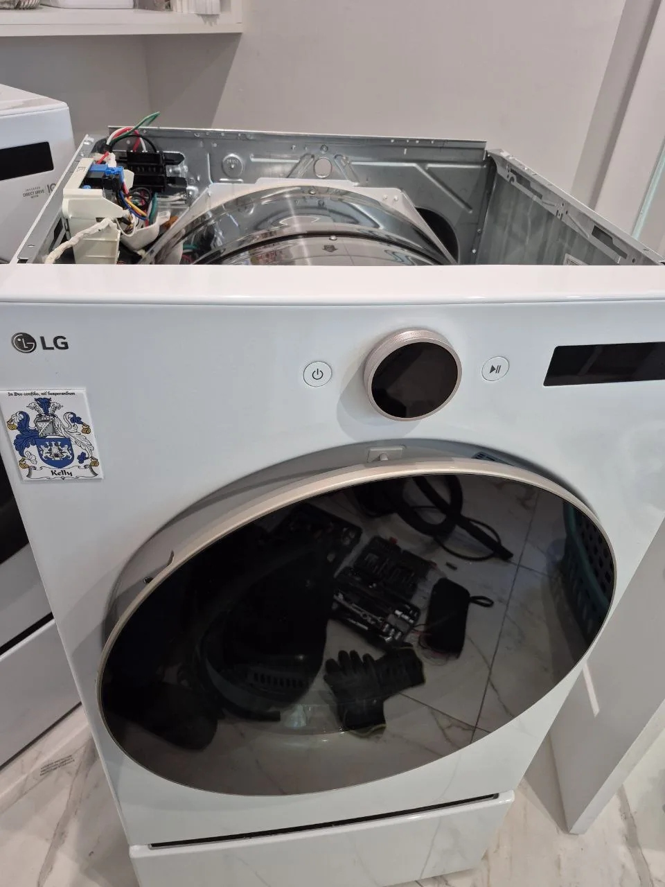A white LG dryer with its top panel removed, revealing internal components and a drum, with repair tools reflected in the