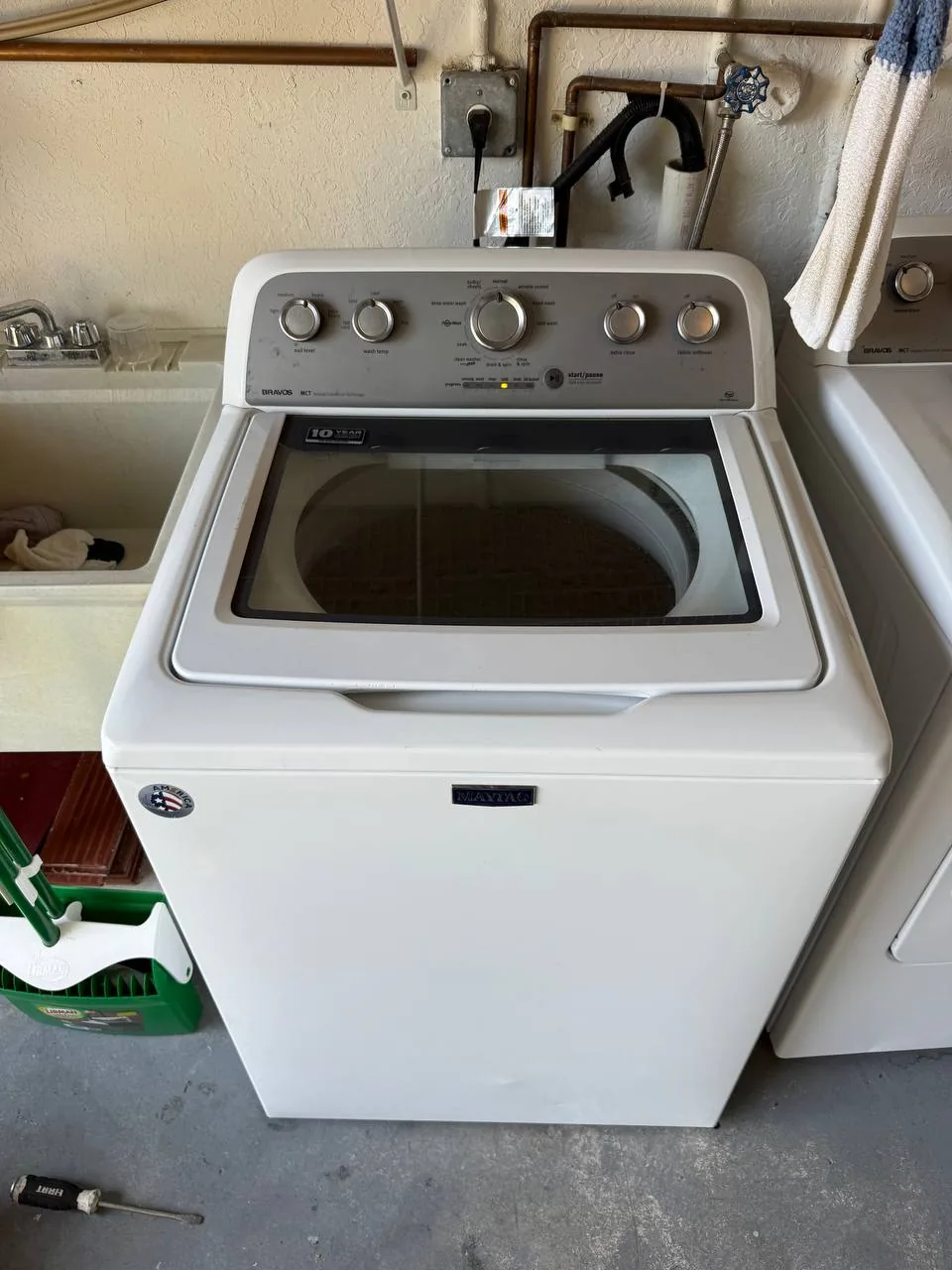 A white Maytag washing machine in a utility room with a sink and tools on the floor