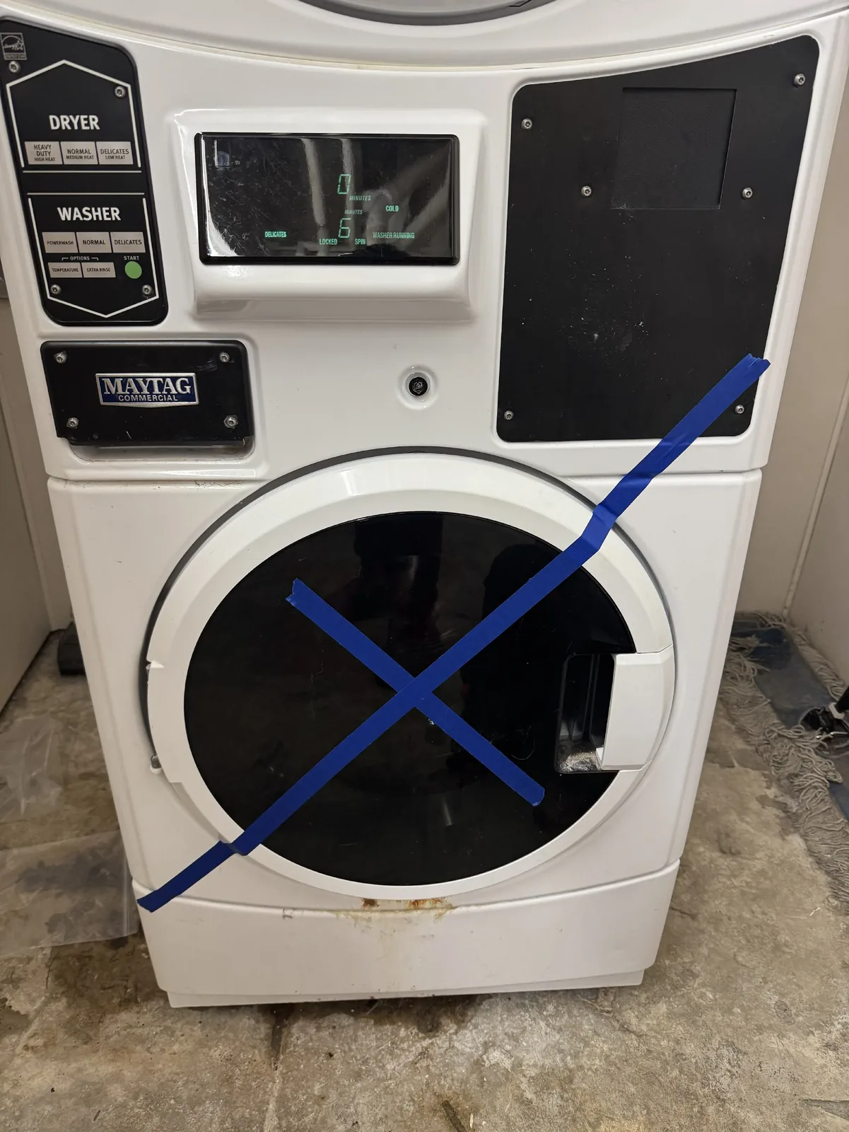 A white Maytag Commercial washer and dryer combo unit with blue tape forming an 'X' on the washer door, indicating it is out