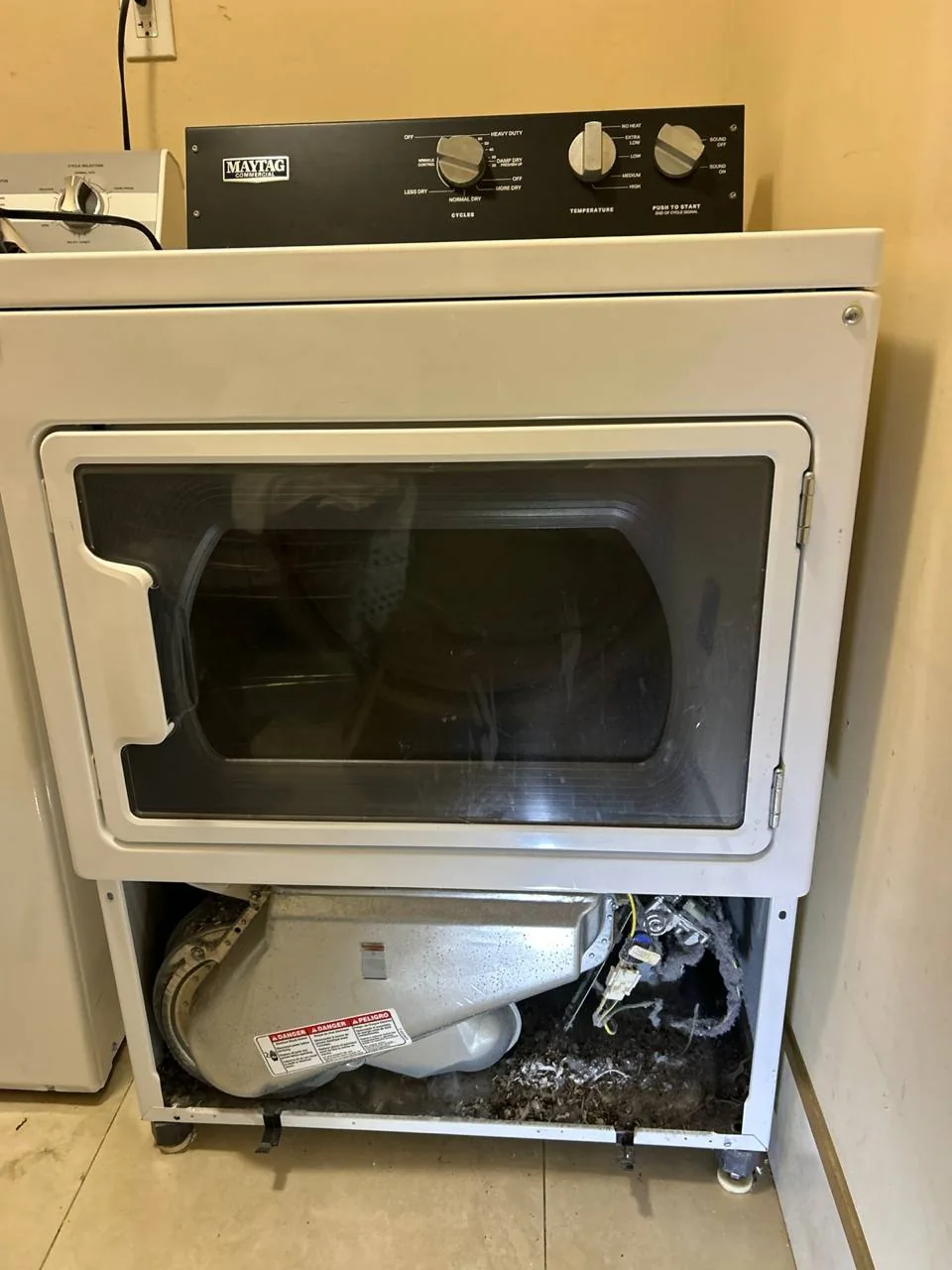 A Maytag dryer with its lower panel removed, showing lint buildup and internal components