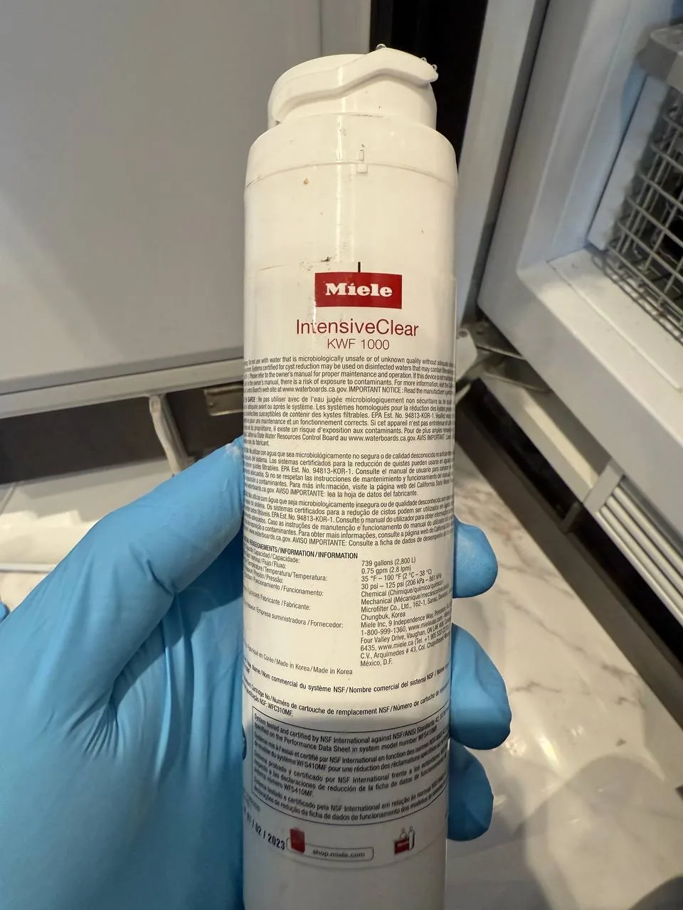 A gloved hand holds a Miele refrigerator water filter, showing product details and a technician working on an appliance