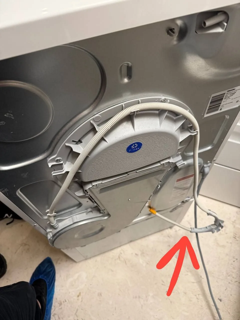 A technician's foot with a blue shoe cover next to the back of a Miele dryer with a red arrow pointing towards a cable