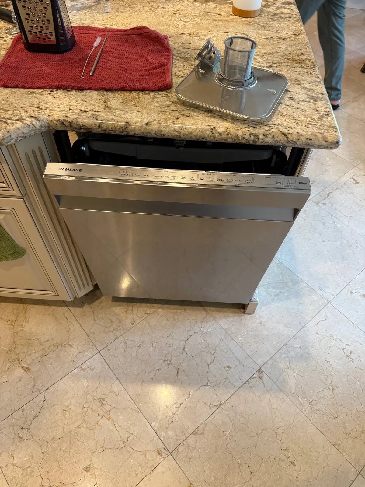 A Samsung dishwasher with its door slightly ajar and a component resting on the granite countertop next to it, along with a