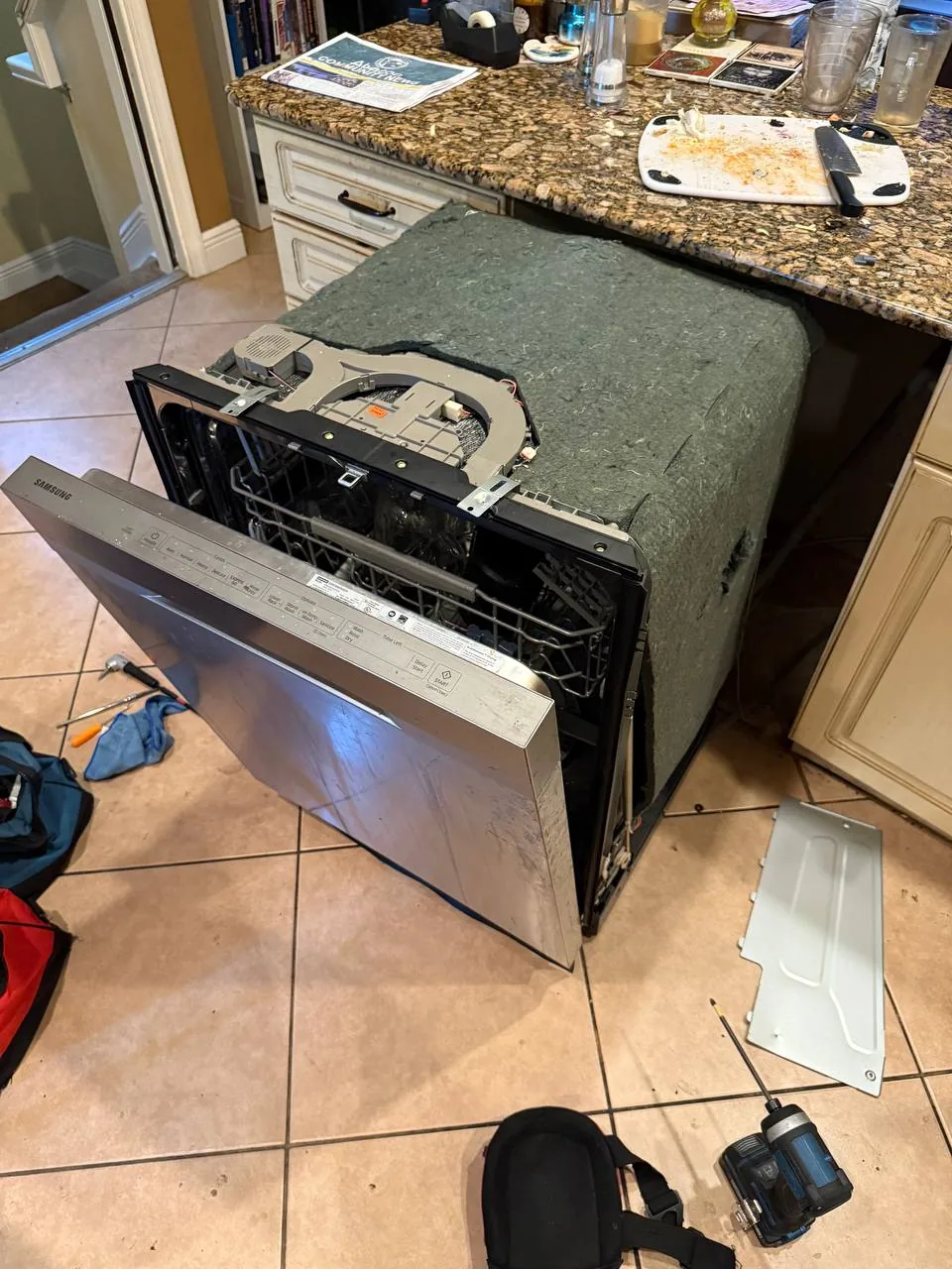 A Samsung dishwasher is pulled out from under the counter, with tools and removed parts around it on the tiled floor