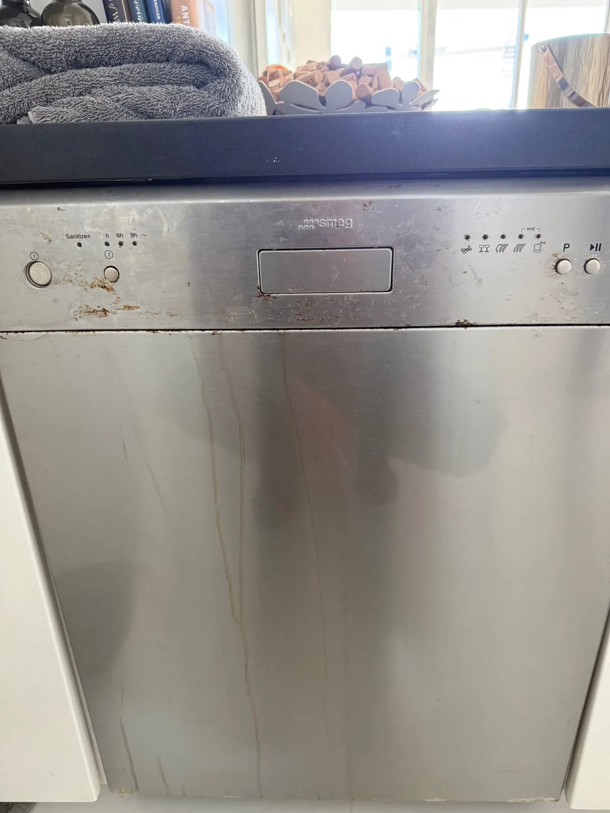 A Smeg dishwasher with a stainless steel finish, showing some rust spots and water marks around the control panel