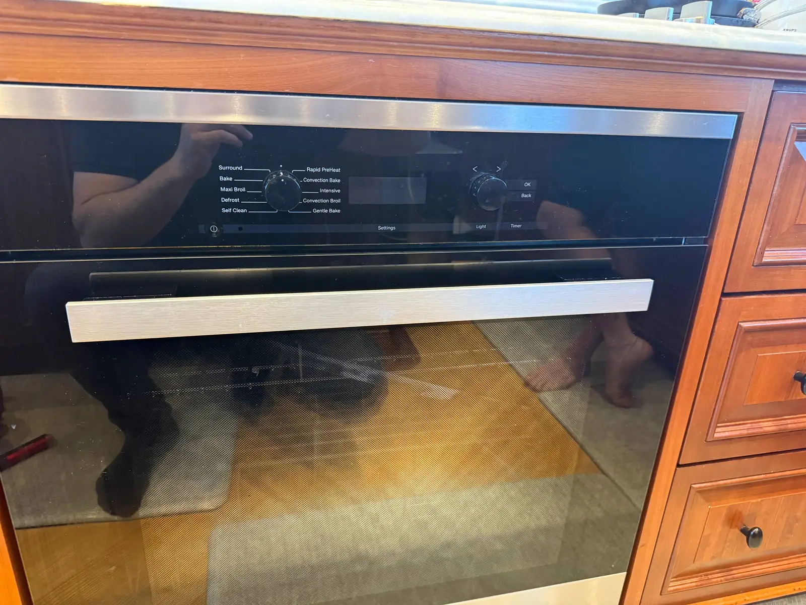 A modern built-in oven with its digital display and control knob visible, reflecting the technician's arms and torso