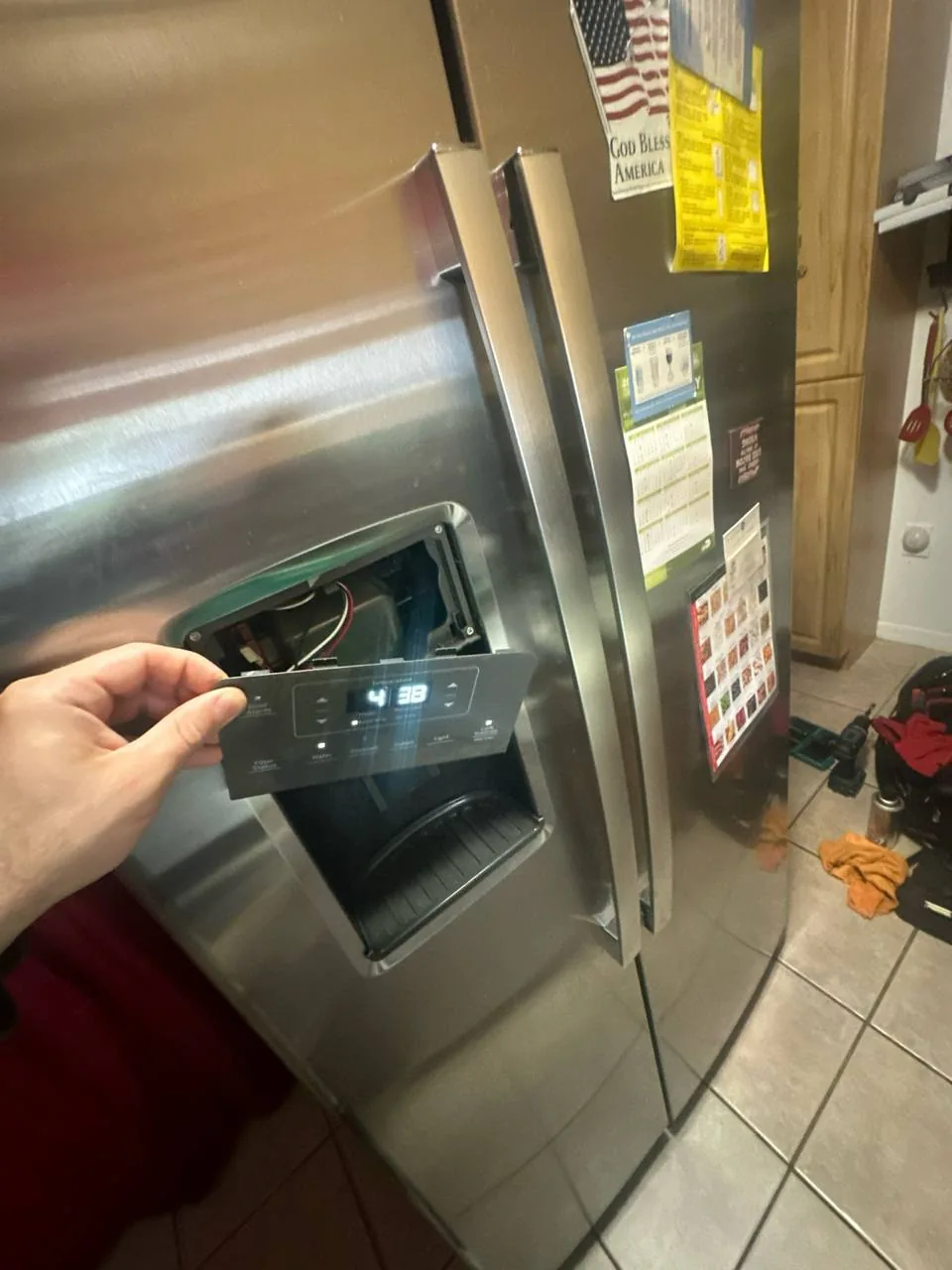 A technician's hand working on the control panel of a stainless steel refrigerator's ice and water dispenser