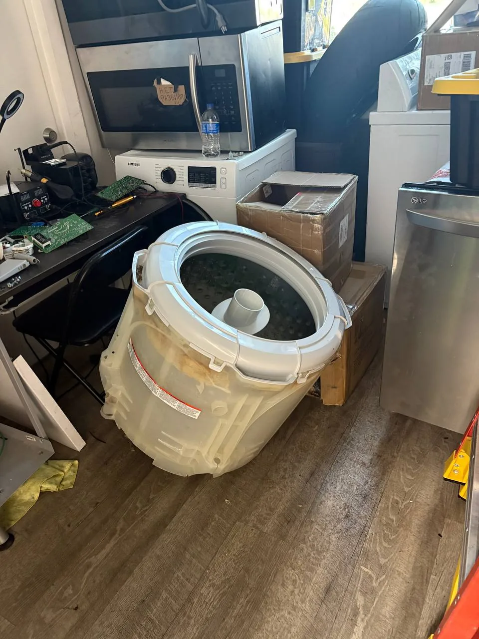 A disassembled washing machine tub is on the floor with other appliances and repair parts in the background