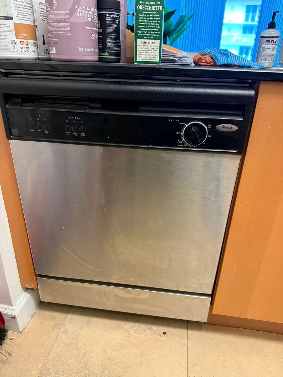 A stainless steel Whirlpool dishwasher installed under a kitchen counter, with household items on the counter above it