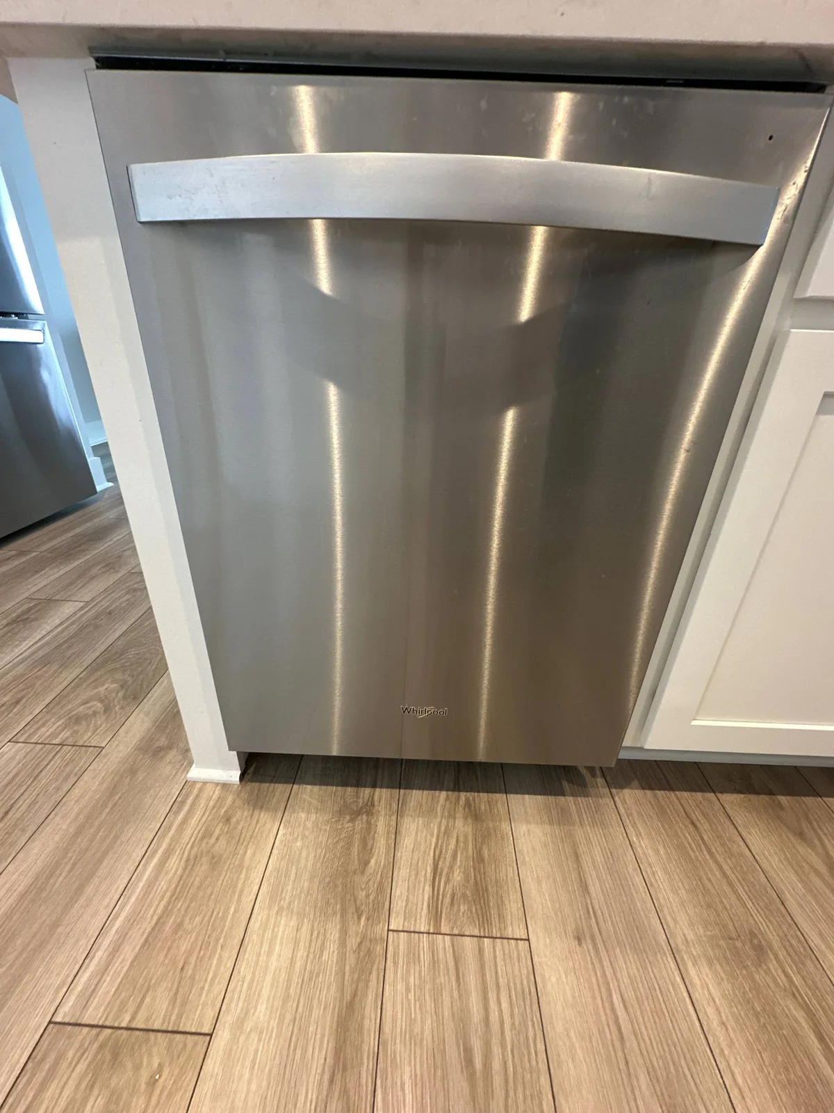 A stainless steel Whirlpool dishwasher is installed between white cabinets above a light wood floor