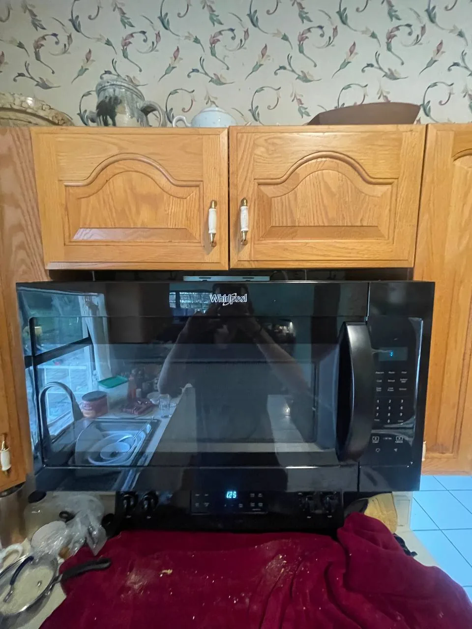 A technician is reflected in a black Whirlpool microwave oven installed above a range with a red cloth and debris on the