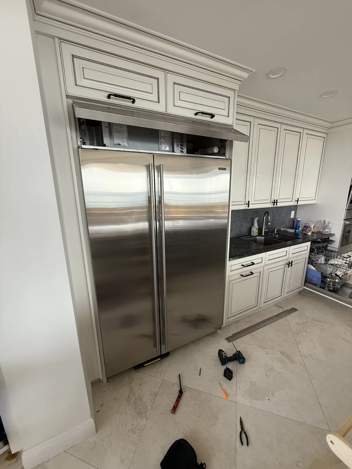 A stainless steel refrigerator with its top panel open, exposing internal components, and repair tools are scattered on the