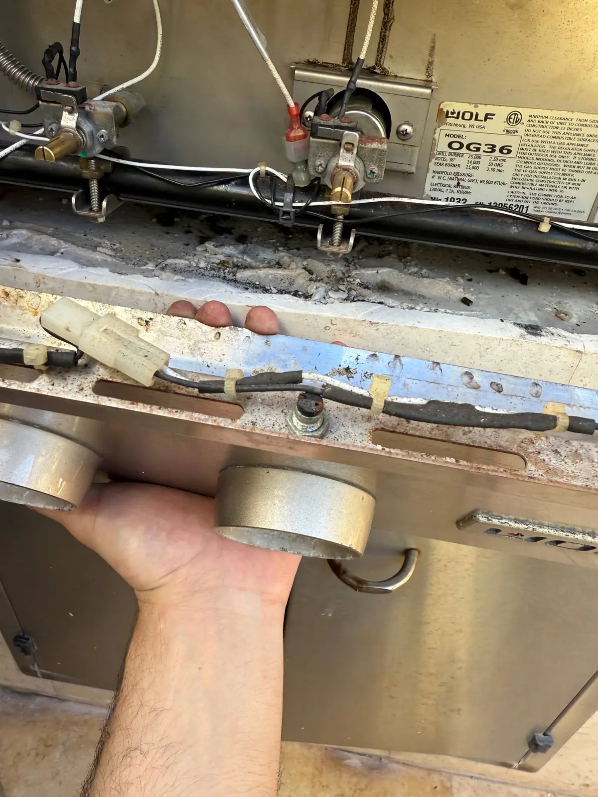 A technician's hand working on the internal components and wiring of a Wolf outdoor cooking appliance